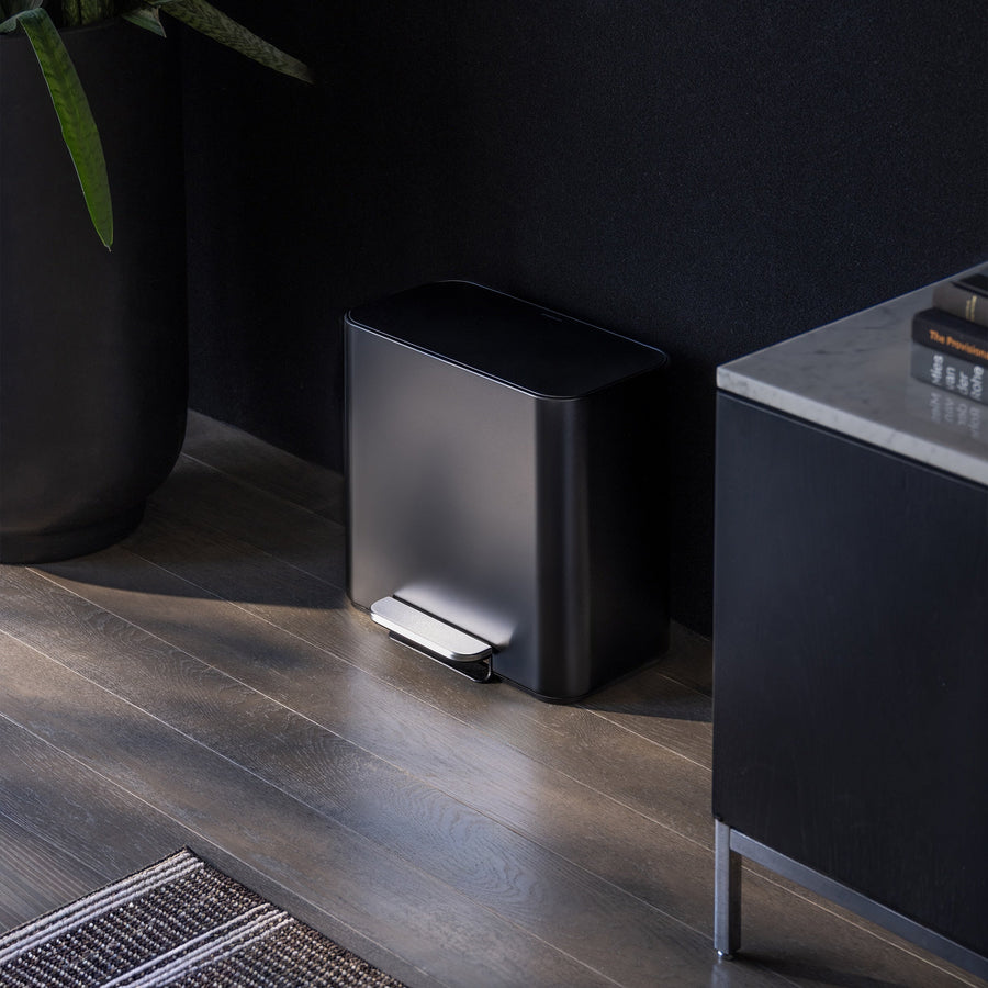 Simplehuman 16L Matte Black Mini Step Can with foot pedal, positioned on wood flooring next to a black storage cabinet with books next to it and plants on the other side.
