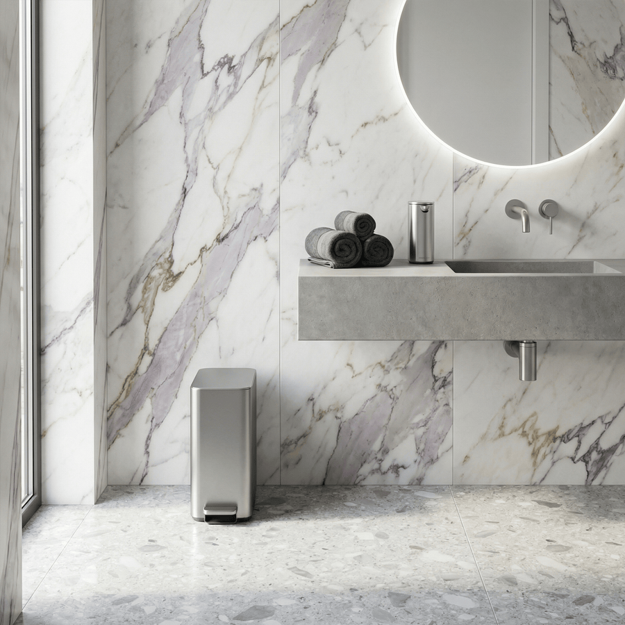 Simplehuman's 9L Slim Step Can in Brushed in a modern, clean bathroom setting featuring a marble wall, bring mirror, and silver appliances. There is also a Brushed Simplehuman Sensor Pump on the sink counter.