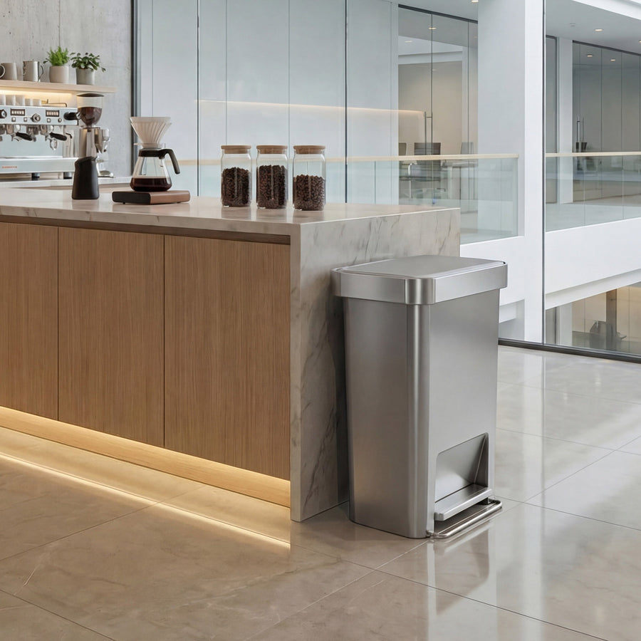 Simplehuman's 45L Hybrid Step Can in Alloy in an office coffee area setting. It is placed against a well-lit marble counter top with wooden drawers. 