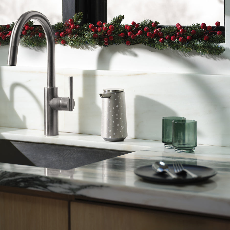 Modern kitchen sink with stainless steel appliances, marble countertop, and an angle view of the Simplehuman Brushed 10 oz. Sensor Pump Foam, decorated with a Simplehuman 2025 Holiday Sleeve. Green glass cups and festive greenery with red berries line the window.