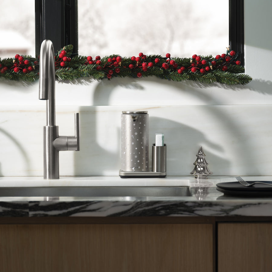 Modern kitchen sink with stainless steel appliances, marble countertop, and a front view Simplehuman Brushed 14 oz. Liquid Sensor Pump, decorated with a Simplehuman 2025 Holiday Sleeve. Green glass cups and festive greenery with red berries line the window.
