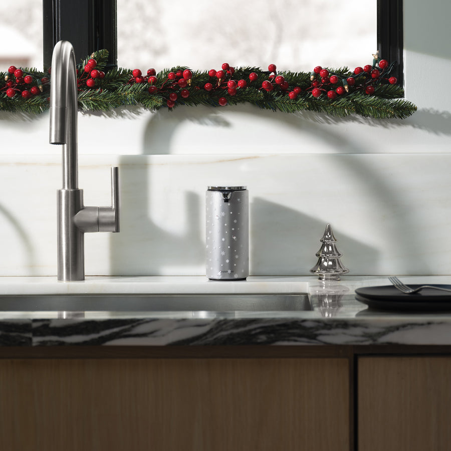 Modern kitchen sink with stainless steel appliances, marble countertop, and a front view of the Simplehuman Brushed 9 oz. Liquid Sensor Pump, decorated with a Simplehuman 2025 Holiday Sleeve. Green glass cups and festive greenery with red berries line the window.