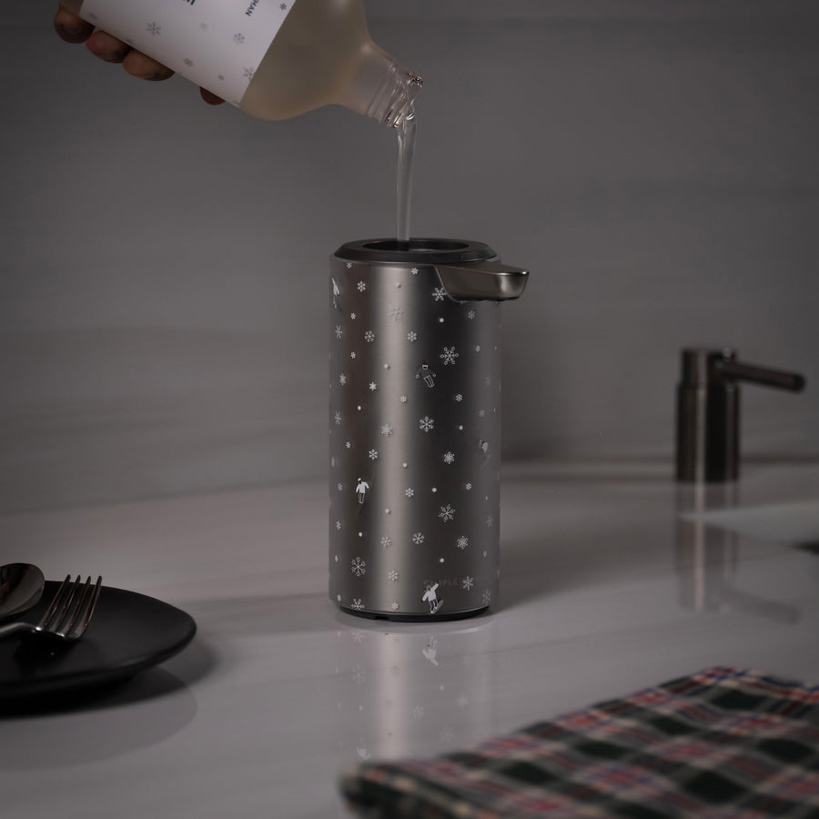 Person pouring Simplehuman's 2025 Holiday Soap in Fresh Pine into their 9 oz. Liquid Sensor Pump that has a 2025 Limited Edition Holiday Sleeve with skiers and snowflakes on a counter top. The counter also features a plaid towel as well as a plate with a spoon and fork on top.