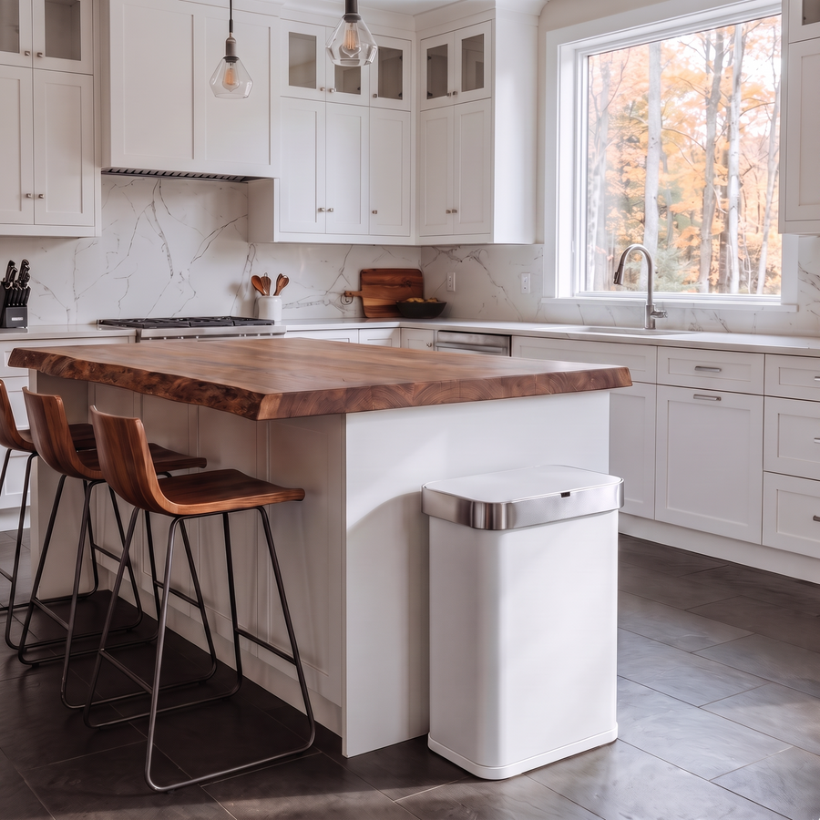 Simplehuman's White Sensor Can in a modern kitchen setting with white cabinets and large window with a view of trees. 