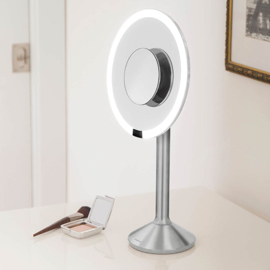Lifestyle view of Simplehuman Sensor Mirror Pro Round, Brushed Stainless Steel, tru-lux light on vanity with makeup.